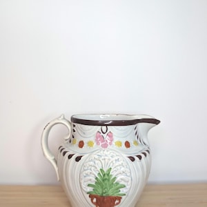 May include: White ceramic pitcher with a brown rim and floral design. The pitcher has a green plant with white flowers in a brown pot. The flowers are pink, yellow, and white.