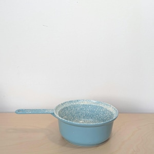 May include: A light blue enamel saucepan with a long handle. The rim and interior have a speckled white pattern. The pan sits on a light wood surface against a white wall. This vintage-style cookware is ideal for cooking.