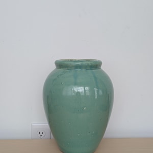 May include: A large, turquoise-colored ceramic vase with a rounded body and a slightly flared rim. The vase has a glossy finish and is displayed on a light-colored wooden surface against a white wall. The vase is a decorative item.