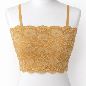 May include: A mustard yellow lace crop top featuring a floral pattern, a straight neckline, and thin shoulder straps. The garment has a scalloped edge along the top and bottom hem, showcasing a detailed lace texture throughout the entire piece.