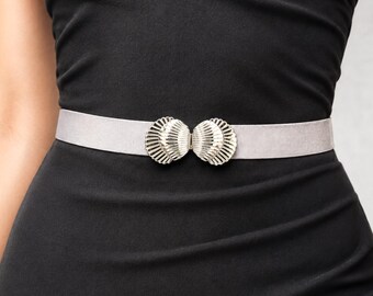 May include: A silver belt with a decorative buckle. The buckle has two shell-shaped elements. The belt is worn over a black dress, creating a contrast between the light belt and the dark dress. The belt is approximately 3 cm wide.