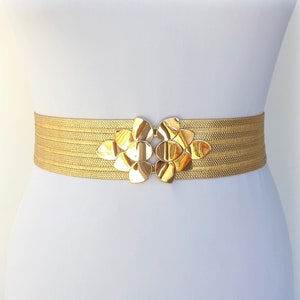 May include: A gold-coloured elastic belt with a large, ornate, gold-toned buckle. The buckle is made of multiple overlapping, rounded, petal-like shapes.