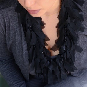May include: A black fabric necklace with a layered, fringed design. The necklace is worn over a grey top. The necklace has a beaded accent. The woman is wearing a grey top.