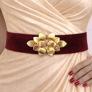 Puede incluir: Un cintur&oacute;n de terciopelo color burdeos intenso con una gran hebilla de metal dorado pulido en forma de flor estilizada con p&eacute;talos redondeados superpuestos. El cintur&oacute;n se lleva sobre un vestido color champ&aacute;n claro con corpi&ntilde;o drapeado.