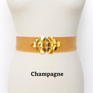 Puede incluir: Un cintur&oacute;n de terciopelo color champ&aacute;n con una hebilla central de metal dorado con un dise&ntilde;o sim&eacute;trico de p&eacute;talos superpuestos. El cintur&oacute;n se muestra sobre un torso de maniqu&iacute;, con la palabra Champagne impresa en texto negro debajo.
