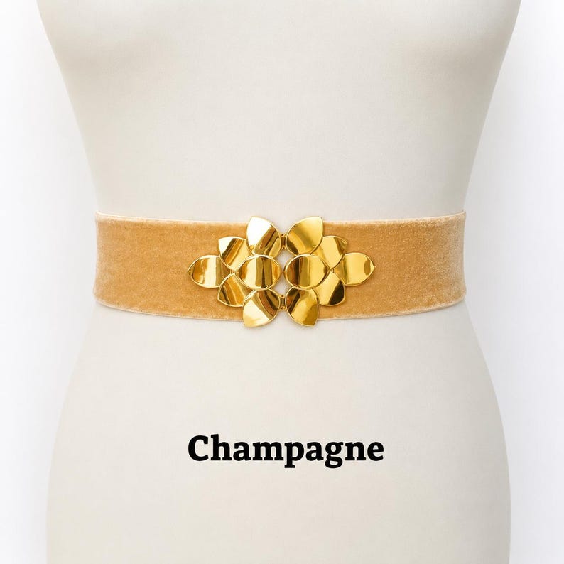 Puede incluir: Un cintur&oacute;n de terciopelo color champ&aacute;n con una hebilla central de metal dorado con un dise&ntilde;o sim&eacute;trico de p&eacute;talos superpuestos. El cintur&oacute;n se muestra sobre un torso de maniqu&iacute;, con la palabra Champagne impresa en texto negro debajo.
