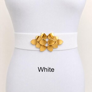 Puede incluir: Un cintur&oacute;n el&aacute;stico blanco con una hebilla central de metal dorado en forma de racimo de p&eacute;talos superpuestos. La palabra White est&aacute; impresa en texto negro debajo del cintur&oacute;n sobre el fondo liso.