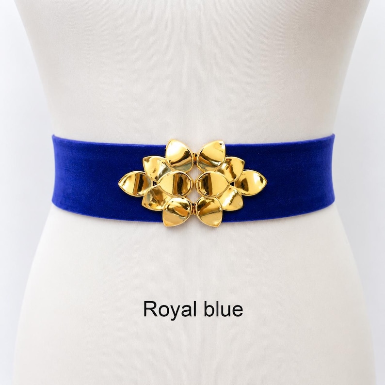 Puede incluir: Un cintur&oacute;n el&aacute;stico azul real con un cierre central de metal dorado en forma de racimo de hojas lisas y redondeadas. El cintur&oacute;n tiene una textura suave similar al terciopelo y se muestra sobre una forma neutra con el texto Royal blue debajo.