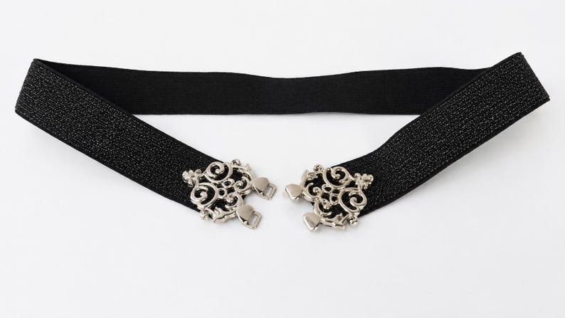 Pu&ograve; includere: Una cintura elastica nera con una superficie testurizzata scintillante e due chiusure in metallo color argento. Le chiusure presentano intricati motivi a volute simmetriche e sono progettate per agganciarsi al centro della vita.