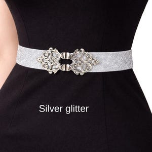 Pu&ograve; includere: Una cintura elastica glitterata color argento con una fibbia decorativa in metallo caratterizzata da intricati motivi a volute. La cintura &egrave; posizionata orizzontalmente su un capo nero a tinta unita. Il testo "Silver glitter" &egrave; stampato sotto.