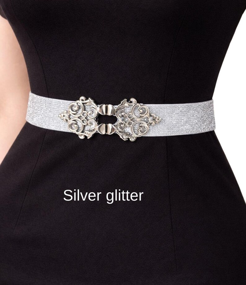 Pu&ograve; includere: Una cintura elastica glitterata color argento con una fibbia decorativa in metallo caratterizzata da intricati motivi a volute. La cintura &egrave; posizionata orizzontalmente su un capo nero a tinta unita. Il testo "Silver glitter" &egrave; stampato sotto.