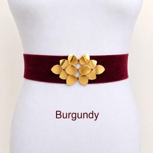 Puede incluir: Un cintur&oacute;n de terciopelo color burdeos con una hebilla central de metal dorado en forma de flor estilizada con varios p&eacute;talos redondeados. La palabra Burgundy est&aacute; impresa en texto rojo oscuro debajo del cintur&oacute;n sobre fondo blanco.