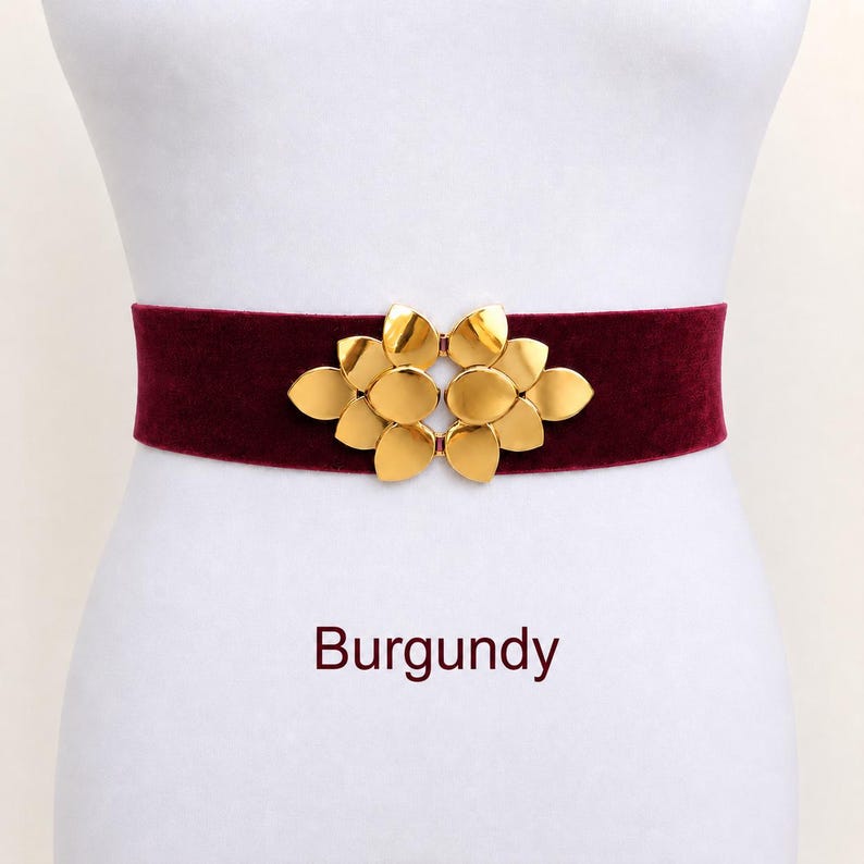 Puede incluir: Un cintur&oacute;n de terciopelo color burdeos con una hebilla central de metal dorado en forma de flor estilizada con varios p&eacute;talos redondeados. La palabra Burgundy est&aacute; impresa en texto rojo oscuro debajo del cintur&oacute;n sobre fondo blanco.