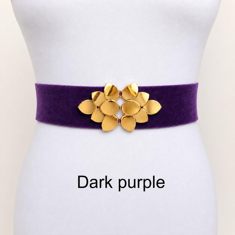 Puede incluir: Un cintur&oacute;n de terciopelo morado oscuro con una hebilla central de metal dorado en forma de racimo de p&eacute;talos estilizados. El cintur&oacute;n tiene un dise&ntilde;o de banda ancha y lisa, e incluye el texto Dark purple impreso debajo sobre el fondo blanco.
