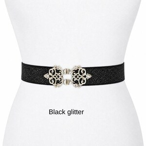 Pu&ograve; includere: Una cintura nera glitterata con una chiusura in metallo color argento caratterizzata da un design ornamentale a volute simmetriche. La cintura presenta una superficie testurizzata e scintillante, fissata al centro da una fibbia decorativa. Testo: Black glitter.