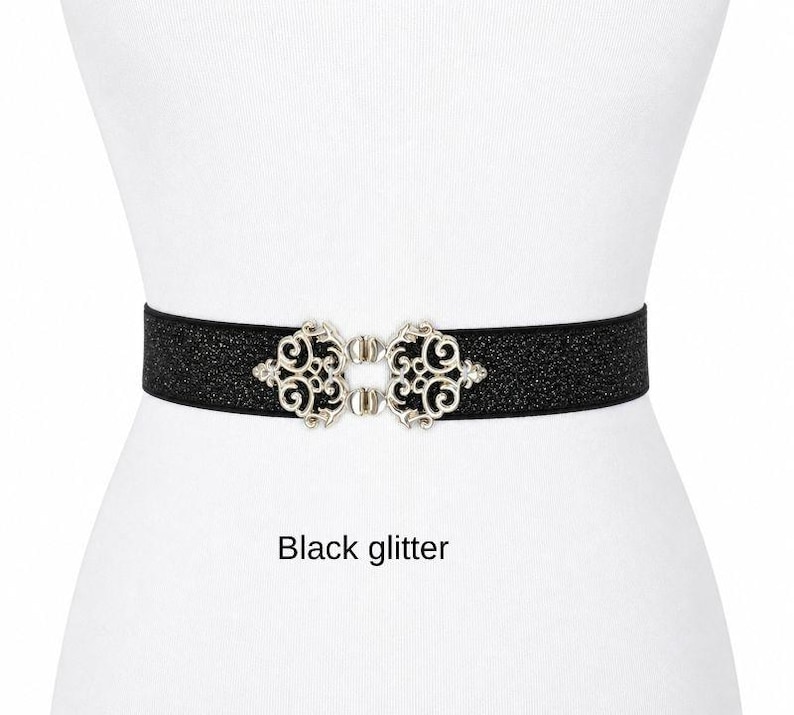 Pu&ograve; includere: Una cintura nera glitterata con una chiusura in metallo color argento caratterizzata da un design ornamentale a volute simmetriche. La cintura presenta una superficie testurizzata e scintillante, fissata al centro da una fibbia decorativa. Testo: Black glitter.
