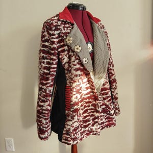 May include: A textured blazer in a red, white, and black pattern, displayed on a mannequin. The blazer features a red collar, a gray and white patterned lapel, and decorative buttons. The lining is black with a red stripe detail.