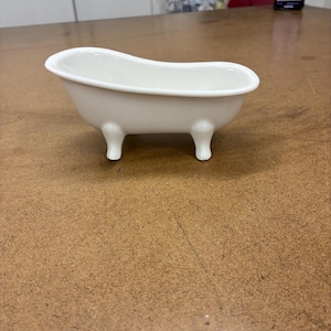 Könnte beinhalten: Eine kleine, weiße Keramikbadewanne mit vier abgerundeten Füßen. Die Miniaturwanne hat eine klassische, ovale Form und ist für dekorative Zwecke konzipiert. Die glänzende Oberfläche reflektiert das Licht und unterstreicht ihre klaren Linien und das schlichte Design. Dieser Artikel eignet sich für die Inneneinrichtung.
