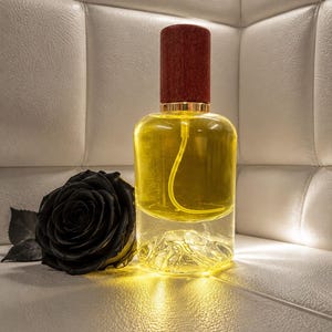 May include: A clear glass perfume bottle filled with yellow liquid, featuring a dark red wooden cap and a molded mountain-shaped base. A single black rose with visible petals and leaves is positioned directly next to the bottle.