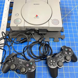 May include: A gray PlayStation console with two black controllers. The console has the Sony logo and PlayStation symbol. The controllers have buttons and directional pads. The console is on a blue grid surface.
