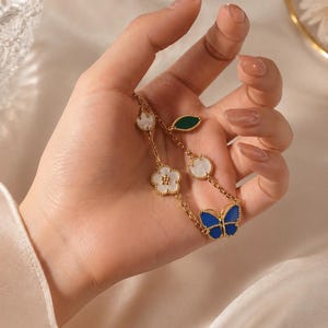 May include: Gold-toned necklace featuring a butterfly and floral design. The necklace has a blue butterfly pendant, white flower and leaf accents. The jewelry is held in a hand against a soft, white background.