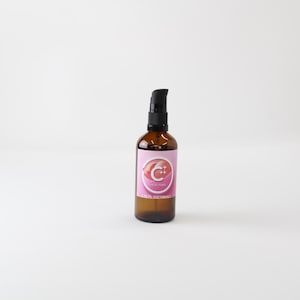 Op de afbeelding: Amberkleurige glazen fles met een zwarte pompdosator. De fles heeft een roze etiket met een wit en rood ontwerp. Waarschijnlijk een huidverzorgings- of cosmetisch product.
