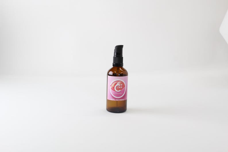 Op de afbeelding: Amberkleurige glazen fles met een zwarte pompdosator. De fles heeft een roze etiket met een wit en rood ontwerp. Waarschijnlijk een huidverzorgings- of cosmetisch product.