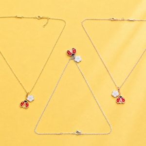 May include: Three necklaces with thin gold-toned chains. Each necklace features a pendant consisting of a small red and black enamel ladybug charm paired with a white five-petaled flower charm. The necklaces are arranged on a bright yellow background.