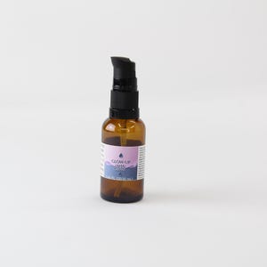 Op de afbeelding: Een kleine bruine glazen fles met een zwarte pompdosator. De fles heeft een etiket met de tekst "GLOW UP AHA CREAM". Het etiket heeft een paars en blauw ontwerp. De fles staat op een witte achtergrond.