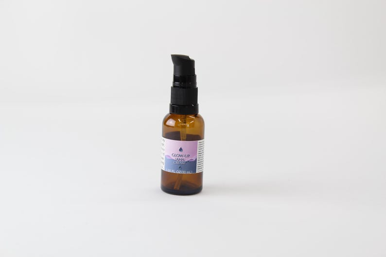 Op de afbeelding: Een kleine bruine glazen fles met een zwarte pompdosator. De fles heeft een etiket met de tekst "GLOW UP AHA CREAM". Het etiket heeft een paars en blauw ontwerp. De fles staat op een witte achtergrond.