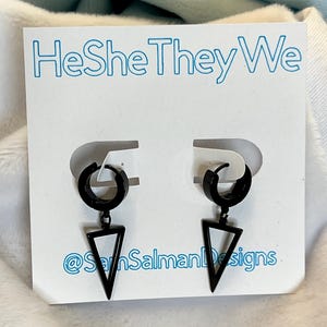 Puede incluir: Un par de pendientes de aro negros con un colgante triangular. Se muestran en una tarjeta blanca con el texto azul "HeSheTheyWe" y el nombre de usuario de Instagram "@SamSalmanDesigns".