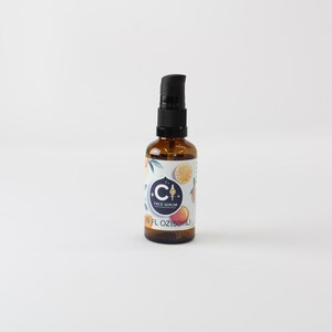 Op de afbeelding: Een bruine glazen fles met een zwarte pompdosator. Het etiket heeft een blauwe cirkel met de letter "C" en de woorden "FACE SERUM". Het etiket heeft ook illustraties van citrusvruchten. De fles bevat 50 ml.