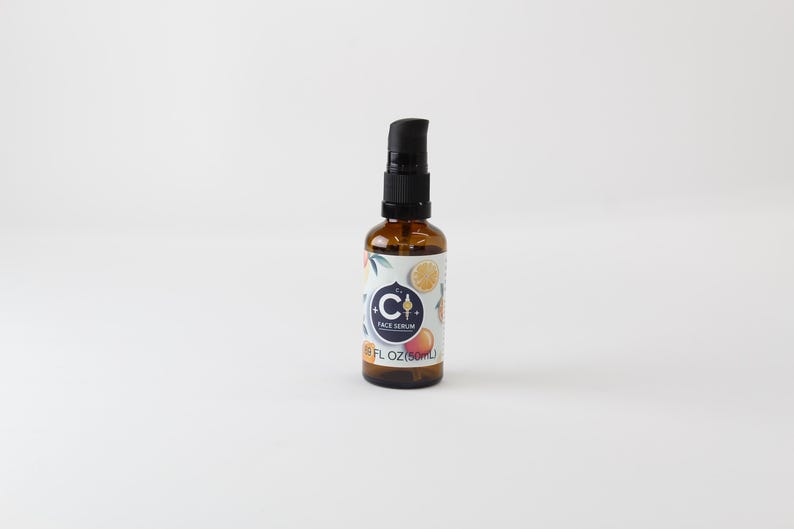 Op de afbeelding: Een bruine glazen fles met een zwarte pompdosator. Het etiket heeft een blauwe cirkel met de letter "C" en de woorden "FACE SERUM". Het etiket heeft ook illustraties van citrusvruchten. De fles bevat 50 ml.