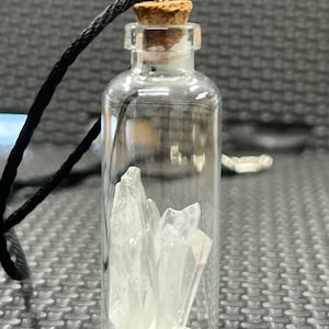 May include: A small, clear glass bottle with a cork stopper, containing several clear quartz crystals. A black cord is attached to the bottle's top. The crystals are clustered inside the bottle, creating a visually appealing display.