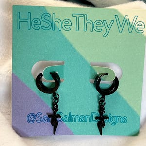 Może przedstawiać: Czarny kolczyki z motywem półksiężyca i zawieszką w kształcie błyskawicy. Kolczyki są prezentowane na turkusowo-fioletowej karcie z napisem "HeSheTheyWe" i nazwą konta na Instagramie "@SarSalmanDesigns".