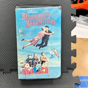 May include: A VHS tape of Disney's "Bedknobs and Broomsticks" features a vibrant cover illustration. The artwork depicts characters floating in a blue underwater scene, with the title text in a playful font. The tape is housed in a black plastic case.