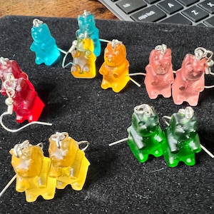 Könnte beinhalten: Eine Sammlung bunter Gummibärchen-Ohrringe in verschiedenen Farben, darunter Rot, Blau, Gelb, Orange, Rosa und Grün. Jeder Ohrring hat einen Gummibärchen-Anhänger mit einem silberfarbenen Haken. Die Ohrringe liegen auf einer schwarzen Oberfläche.