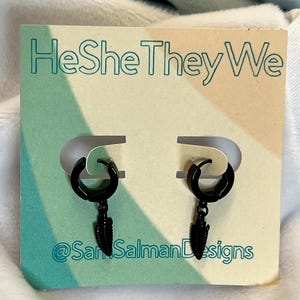 Może przedstawiać: Czarny kolczyki koła z zawieszkami w kształcie piór, prezentowane na karcie z napisem "HeSheTheyWe" i nazwą sklepu, @SamSalmanDesigns. Kolczyki są umieszczone na miękkim, wielokolorowym tle.