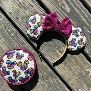 May include: A set of mouse ear headbands and a matching round accessory. The ears and accessory feature a white background with colorful Mickey Mouse silhouettes in leopard and rainbow patterns. A large, sparkly pink bow sits atop the headband.
