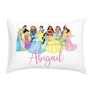 Op de afbeelding: Witte rechthoekige kussensloop met een groep cartoonprinsessen in kleurrijke jurken. De naam "Abigail" is in roze cursief geschreven. De kussensloop is waarschijnlijk gemaakt van zachte stof.