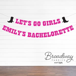 Op de afbeelding: Roze en zwarte banner met de tekst "Let's Go Girls Emily's Bachelorette" en zwarte cowboylaars-silhouetten aan beide uiteinden. De banner hangt aan een witte houten muur. De tekst "Broadway Fabrics and Gifts" staat in zwarte cursieve letters onderaan de afbeelding.