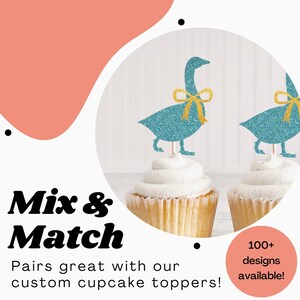 K&ouml;nnte beinhalten: Zwei Cupcakes mit blauen Glitzer-G&auml;nse-Cupcake-Toppern. Die Topper haben goldene Schleifen. Der Text "Mix & Match" ist in fetten schwarzen Buchstaben. Der Text "Pairs great with our custom cupcake toppers!" ist in schwarzen Buchstaben. Der Text "100+ designs available!" ist in wei&szlig;en Buchstaben auf einem rosa Kreis.