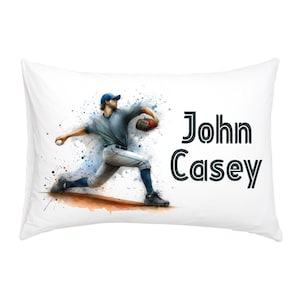 Puede incluir: Funda de almohada blanca con una ilustración de acuarela de un jugador de béisbol lanzando una pelota. El jugador lleva un uniforme azul y blanco. El texto "John Casey" está impreso en negro en la funda de almohada.