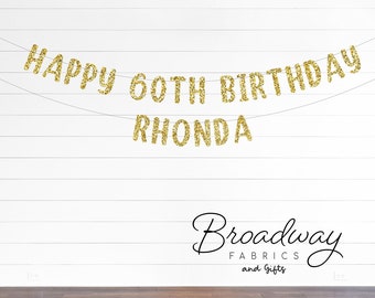 May include: A gold glitter banner spelling out Happy 60th Birthday Rhonda, arranged in two lines on thin strings. The letters have a textured, sparkling finish and are suspended against a white horizontal paneled wall above a dark wood floor.