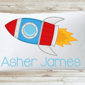 Peut inclure: Une taie d'oreiller blanche avec une illustration de fusée bleue et rouge et le nom "Asher James" imprimé en bleu.