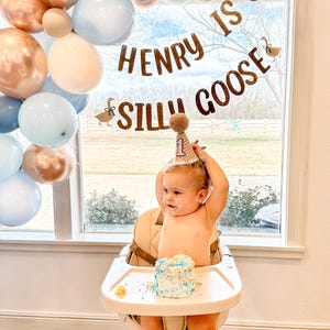 K&ouml;nnte beinhalten: Ein Babyjunge tr&auml;gt eine braune und wei&szlig;e karierte Geburtstags-M&uuml;tze mit einem wei&szlig;en Bommel obenauf und sitzt in einem Hochstuhl mit einem blau-wei&szlig;en, glasierten Kuchen vor sich. Ein Banner hinter dem Baby lautet "Henry is one Silly Goose" mit einer braunen G&auml;nse-Illustration auf jeder Seite. Das Banner ist mit einem blauen, beigen und kupferfarbenen Ballonbogen aufgeh&auml;ngt.