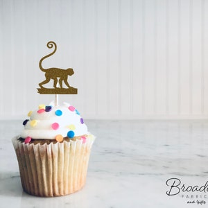 Puede incluir: Un cupcake con glaseado blanco y chispas de colores coronado con un adorno de cupcake de mono de purpurina dorada.
