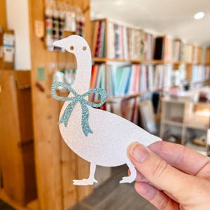 K&ouml;nnte beinhalten: Ein wei&szlig;er Glitzer-G&auml;nse-Ausschnitt mit einer blauen Glitzer-Schleife. Die Gans h&auml;lt eine blaue Glitzer-Schleife im Schnabel.
