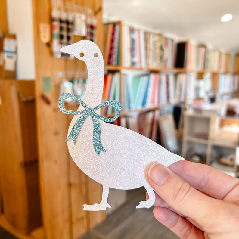 K&ouml;nnte beinhalten: Ein wei&szlig;er Glitzer-G&auml;nse-Ausschnitt mit einer blauen Glitzer-Schleife. Die Gans h&auml;lt eine blaue Glitzer-Schleife im Schnabel.