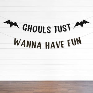 Puede incluir: Una pancarta negra con purpurina que presenta dos siluetas de murciélagos en los extremos y el texto GHOULS JUST WANNA HAVE FUN dispuesto en dos líneas sobre hilos finos. Las letras y los murciélagos tienen un acabado brillante y texturizado.